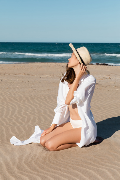 young woman in straw hat sitting on golden sand near blue ocean - Photo, Image