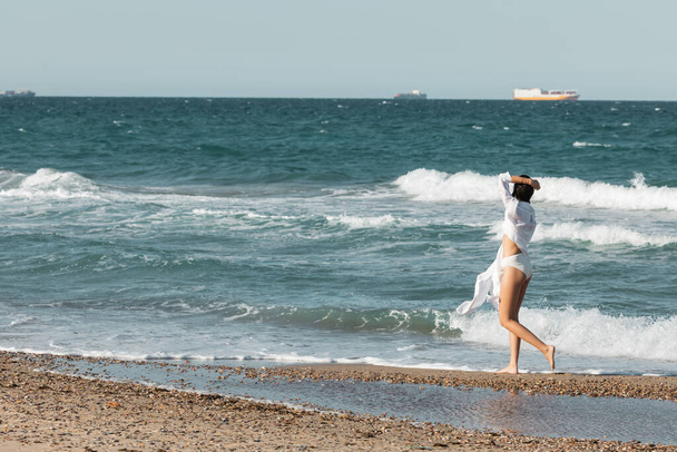 longitud completa de la mujer joven en camisa blanca y trajes de baño caminando en la orilla del mar en la playa  - Foto, imagen