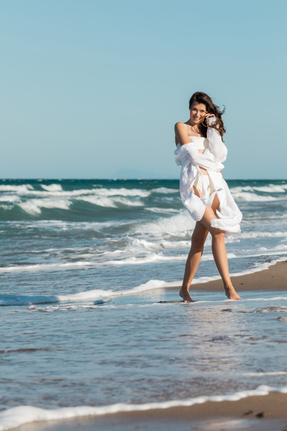 full length of cheerful woman in white shirt and swimwear standing near ocean on beach  - Photo, Image