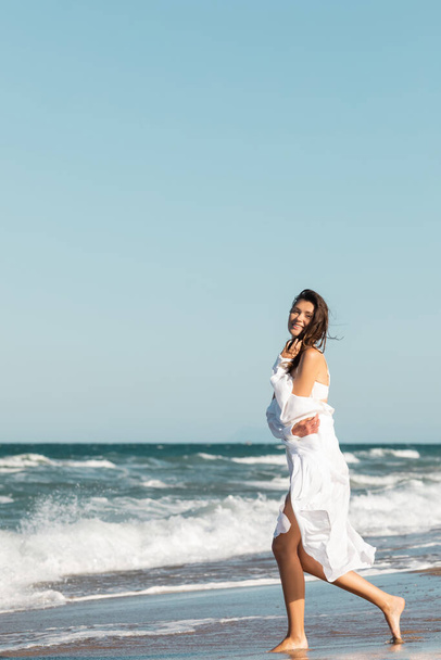 full length of joyful woman in white shirt and swimsuit smiling near ocean on beach - Foto, imagen