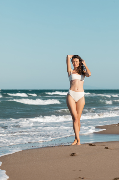 полная длина улыбающейся молодой женщины в белых купальниках, идущей рядом с океаном на пляже - Фото, изображение