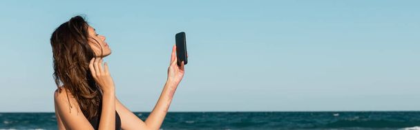 jovem morena em maiô tirando selfie no smartphone perto do mar, banner - Foto, Imagem