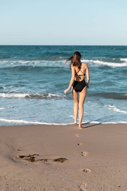 back view of brunette woman in black swimwear walking into sea - Foto, afbeelding