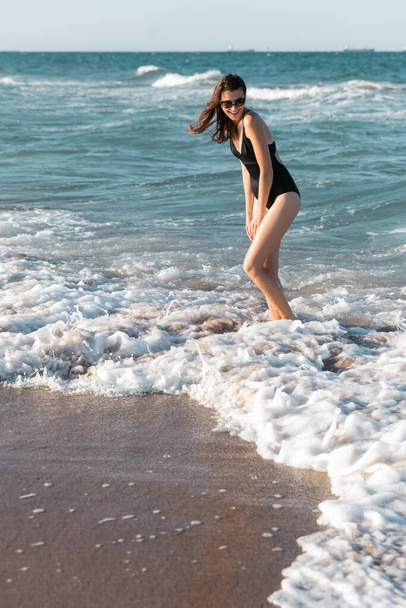 mujer feliz en traje de baño negro y gafas de sol con estilo de pie en el mar - Foto, imagen