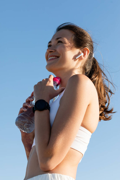 Tiefansicht einer glücklichen Frau im drahtlosen Kopfhörer, die eine Flasche mit Wasser gegen den blauen Himmel hält - Foto, Bild