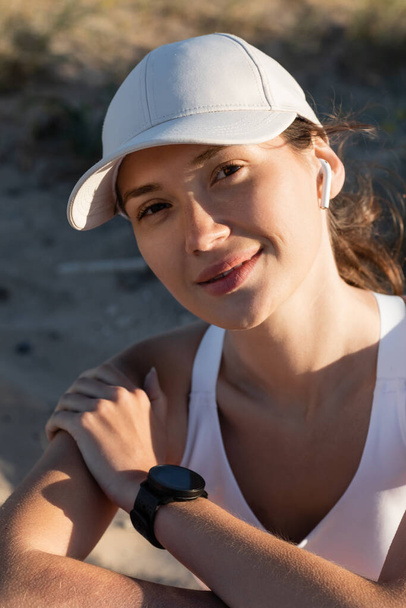 portrait of happy sportswoman in baseball cap listening music in wireless earphone on beach - Fotó, kép