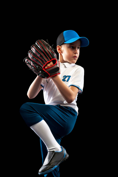 Offensive player. Dynamic portrait of little baseball player, pitcher in blue-white uniform training isolated on black studio background. Concept of sport, achievements, studying, competition. - Photo, image