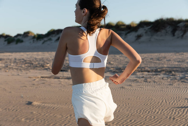 joven morena mujer en ropa deportiva blanca trotando en la playa en verano - Foto, Imagen