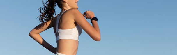 cropped view of cheerful young and sportive woman jogging against blue sky, banner - Photo, Image