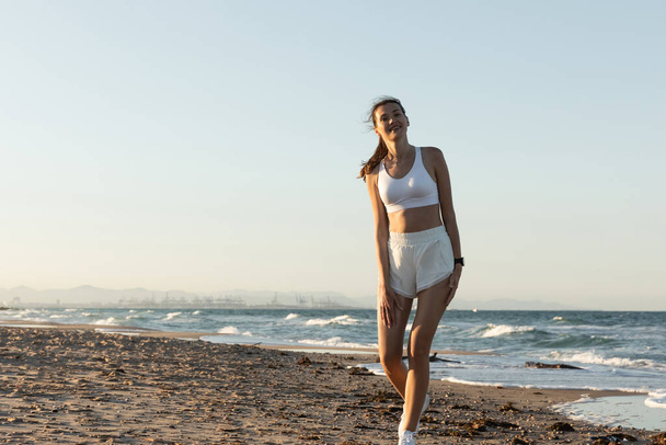 happy woman in white sports bra and shorts standing near sea on beach - Photo, Image