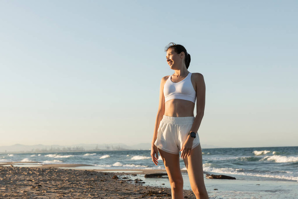 mujer alegre en sujetador deportivo blanco y pantalones cortos de pie cerca del mar en la playa - Foto, imagen