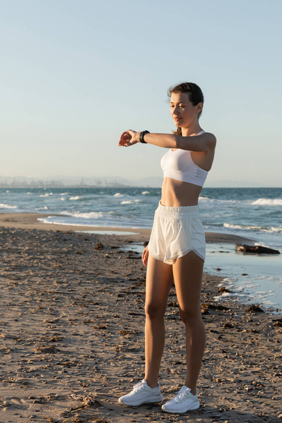 fiatal nő fehér sportmelltartó és rövidnadrág nézi fitness tracker tenger közelében a strandon - Fotó, kép