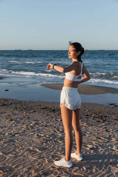 young woman in sports bra and shorts looking at fitness tracker near sea on beach - Photo, Image