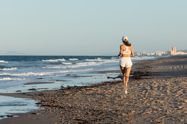 back view of young sportive woman in baseball cap jogging near sea in summer, banner - Foto, Imagem
