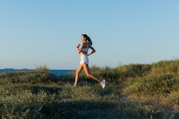 jovem mulher esportiva em shorts e fone de ouvido sem fio correndo na grama perto do mar  - Foto, Imagem