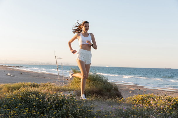 jeune femme en short et écouteurs sans fil jogging sur l'herbe près de la mer - Photo, image