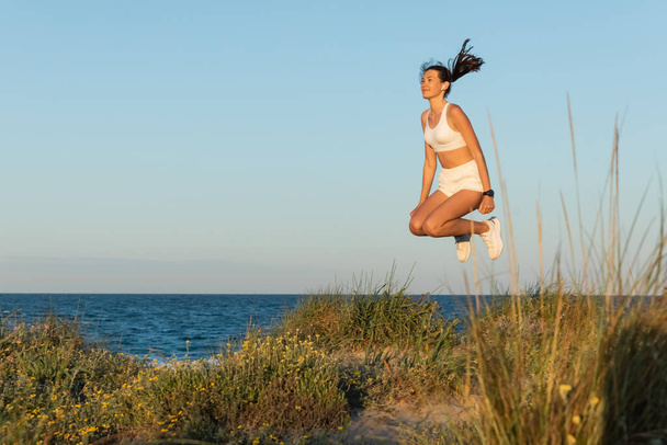 молодая женщина в спортивных шортах и беспроводных наушниках, прыгающая возле синего моря - Фото, изображение