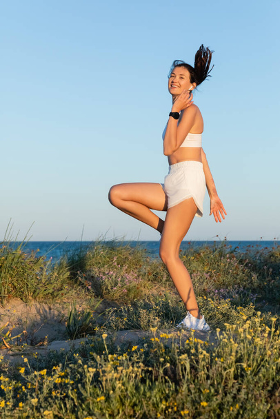 mujer en forma feliz en pantalones cortos deportivos y auriculares inalámbricos saltando cerca del mar azul - Foto, imagen