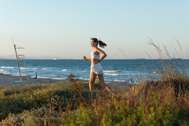 вид сбоку на молодую женщину в спортивном бюстгальтере и беспроводных наушниках, бегущую по траве у берега моря - Фото, изображение