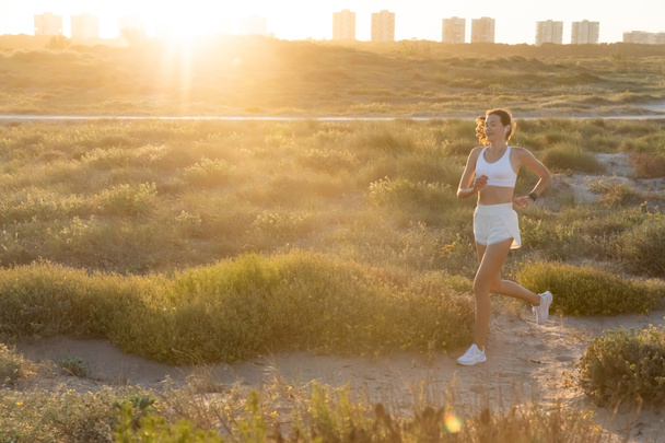 jeune femme sportive dans écouteur sans fil courir pendant le coucher du soleil - Photo, image