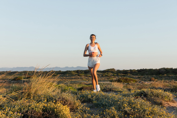 smiling sportswoman in shorts and sports bra listening music and running outside  - Foto, afbeelding