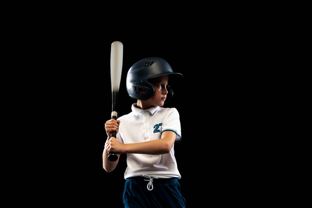 Offensive player. Dynamic portrait of little baseball player, pitcher in blue-white uniform training isolated on black studio background. Concept of sport, achievements, studying, competition. - Foto, imagen