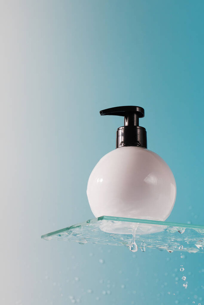 Mockup of a white dispenser with a cosmetic product on glass shelf and falling down drops on blue background closeup. Cosmetic product for body washing - Photo, Image