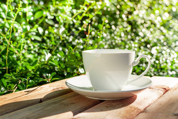weiße Keramik-Kaffeebecher Auf dem Holzboden, grüner Baum Bokeh Hintergrund in der Morgensonne im Garten. Soft focus.flacher Fokus-Effekt. - Foto, Bild
