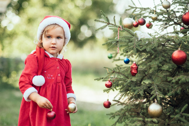 Christmas in july. Child waiting for Christmas in wood in summer. portrait of little girl in red dress decorating christmas tree. winter holidays and people concept. Merry Christmas and Happy Holidays - Fotó, kép