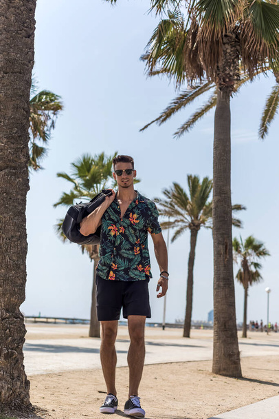 Young man walking on city wearing casual clothes and sunglasses with shorts flowered shirt holding travel bag - Zdjęcie, obraz