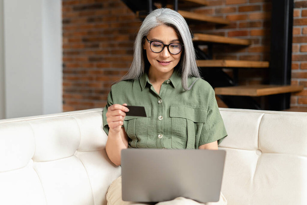 Calm senior businesswoman in glasses sitting at the sofa using laptop and credit card for online payment from home, typing number to enter internet banking account, ordering food - Foto, afbeelding
