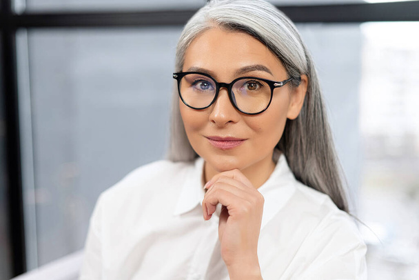 Portrait of senior gray-haired female doctor sitting at the desk in the office, and looking at the camera. Occupation of senior people concept - Zdjęcie, obraz