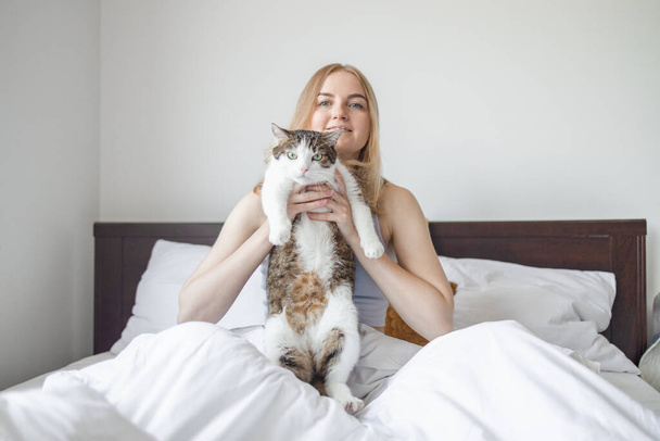 Χαρούμενη ξανθιά τριαντάρα γυναίκα με γκρι δυστυχισμένη γάτα ξαπλωμένη σε λευκό κρεβάτι στο σπίτι το πρωί. Κατοικίδια, άνεση, ξεκούραση και άνθρωποι έννοια.  - Φωτογραφία, εικόνα