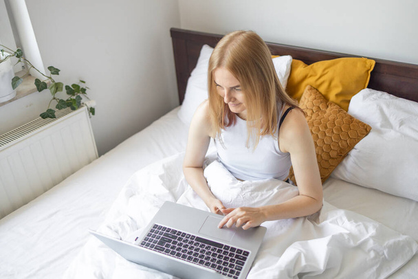 家の中で午前中にベッドに座ってノートパソコンで働く幸せなカジュアルな美しい女性。家庭で働くこと、フリーランスのコンセプト. - 写真・画像