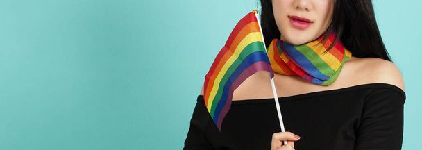 LGBTの女性は、青い緑の背景に立ってプライドフラグを保持します。首に虹色のスカーフを持つアジアのLGBTQの女性。頭のいい明るく元気そうね。LGBTQ多様性の誇りの概念. - 写真・画像