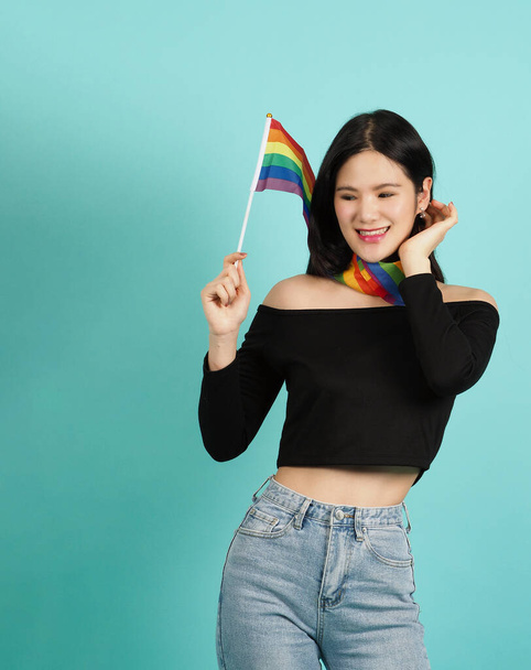 Kobieta LGBTQ z flagą dumy stojąca na niebieskim zielonym tle. Azjatka LGBTQ z tęczowym szalikiem na szyi. wyglądają inteligentne jasne i energiczne wesoły. Koncepcja dumy różnorodności LGBTQ. - Zdjęcie, obraz