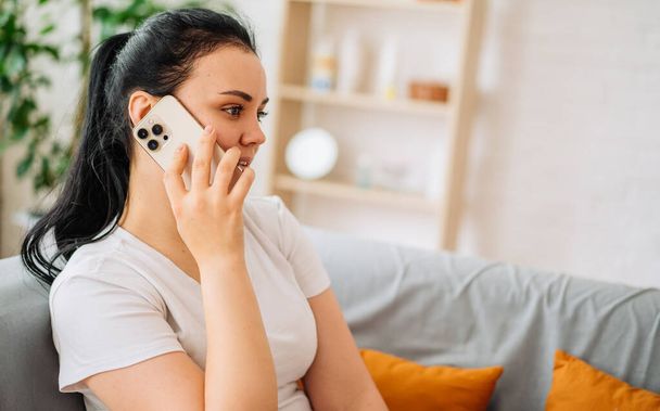 Μια χαμογελαστή γυναίκα μιλάει στο τηλέφωνο στο σπίτι, ένα χαρούμενο νεαρό κορίτσι κρατάει ένα κινητό τηλέφωνο ενώ απαντάει σε μια κλήση - Φωτογραφία, εικόνα