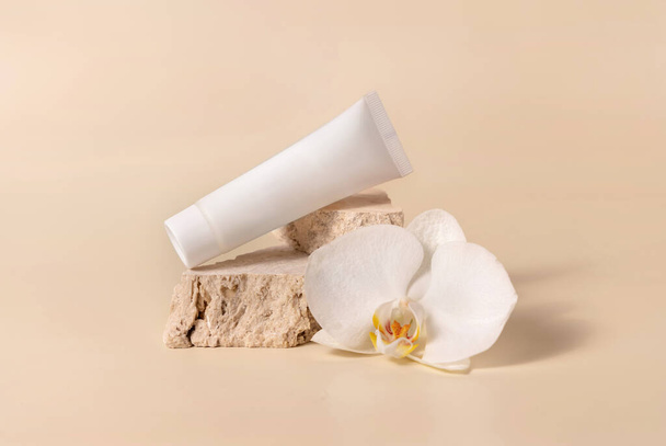 Weiße Cremetubus auf Travertinstein in der Nähe von weißen Orchideenblüten auf hellgelb, Nahaufnahme, Attrappe. Hautpflege handgemachtes Schönheitsprodukt, Creme oder Lotion. Exotische Naturkosmetik - Foto, Bild