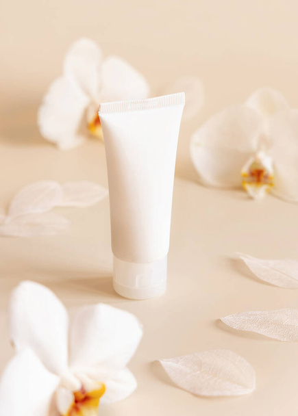 Λευκός πλαστικός σωλήνας κρέμας κοντά σε λευκά λουλούδια ορχιδέας σε ανοιχτό κίτρινο, κοντινό πλάνο, μακιγιαρισμένο. Προϊόν ομορφιάς Skincare, κρέμα ή λοσιόν. Εξωτικά φυσικά καλλυντικά, παστέλ ελάχιστη σύνθεση - Φωτογραφία, εικόνα