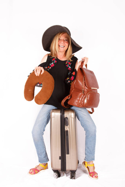 Starší blondýny žena, sedí na kufru připravuje na dovolenou s kloboukem a kabelkou, bílé pozadí - Fotografie, Obrázek