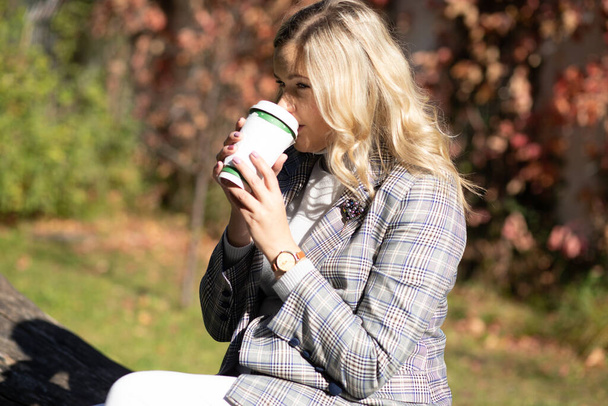 Long-haired blonde 35 years old in tweed jacket drinks hot coffee from recycled cup sitting on autumn day in city park. Ecology and environmental protection. Walking in fresh air. Lifestyle.  - Photo, Image
