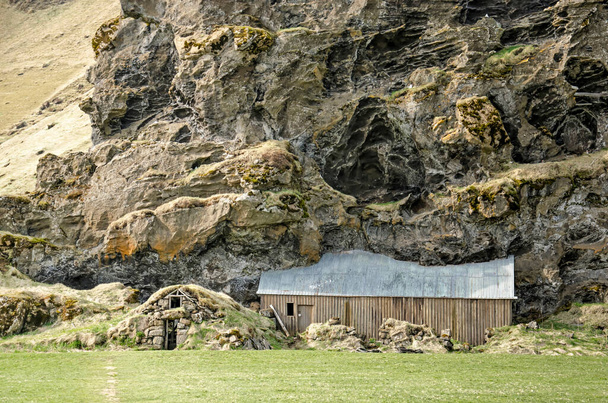 Скогар, Исландия, 22 апреля 2022 года: традиционный дом и сарай, построенные внутри или против большой скалы - Фото, изображение