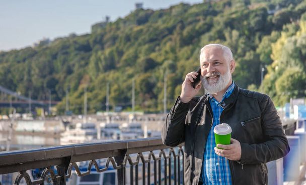 Attractive white bearded man have conversation over smartphone at green part background. Gladsome senior man discussing exciting news over mobile phone while drinking coffee during walking over river pier. - Foto, Imagen