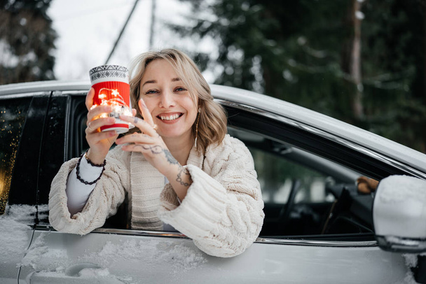 Attractive happy young woman hiding behind the cup of tea looking out the car window. Winter travel. Weekend recreation - 写真・画像