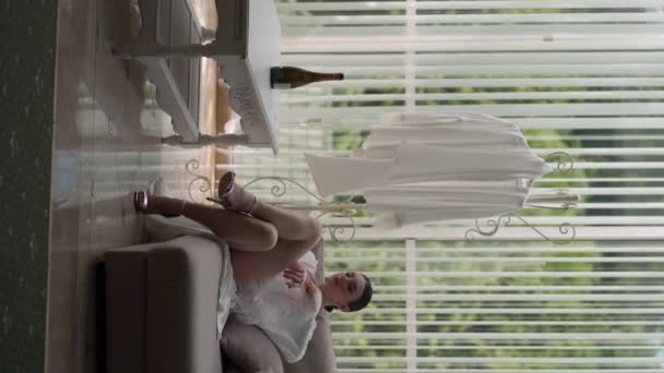 Vertikaalisia videoita. Kaunis yksinäinen nainen valkoisessa takissa on kyllästynyt yksin istumaan sohvalla. Hän on alusvaatteissa. Näyttää tyylikäs ja seksikäs - Materiaali, video