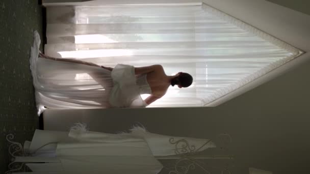 縦型ビデオ。白いバスローブを着た美しい花嫁が窓に立ってポーズをとって - 映像、動画