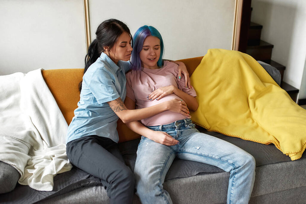 Girl hug and touch belly of pregnant girlfriend partner on sofa at home. Young caucasian lesbain couple spending time together. Homosexual relationship and motherhood. Closeness. Domestic lifestyle - Foto, immagini