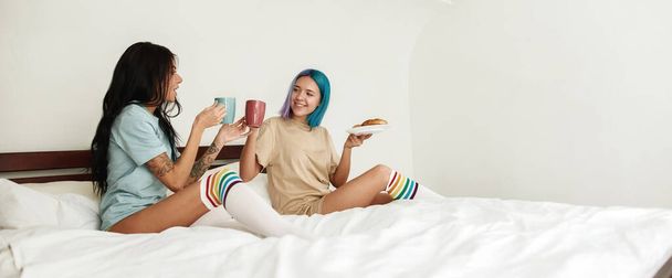 Girl holding plate of buns and drinking tea or coffee with her girlfriend in bed. Young lesbian caucasian couple at home. Homosexual relationship. Domestic lifestyle. Spacious bedroom. Morning time - Foto, imagen