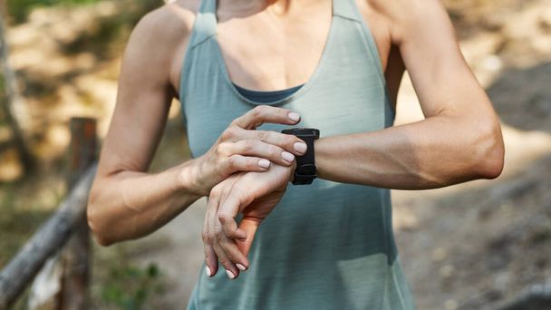 Cropped view of young sportswoman using and browsing smartwatch during sports training outdoors. Slim athletic girl wearing sportswear. Concept of modern healthy lifestyle. Warm summer day - Foto, Imagen