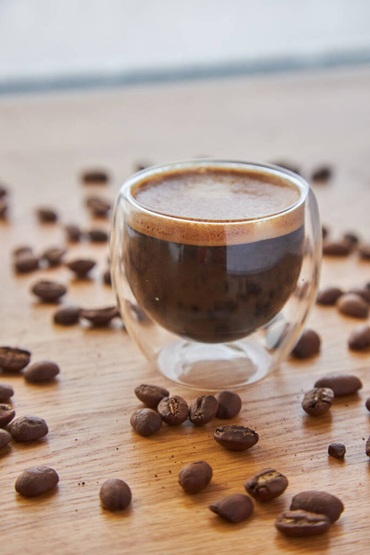 Espresso in einer transparenten Tasse, die auf einer hölzernen Tischplatte steht, umgeben von Kaffeebohnen im Hintergrund - Foto, Bild